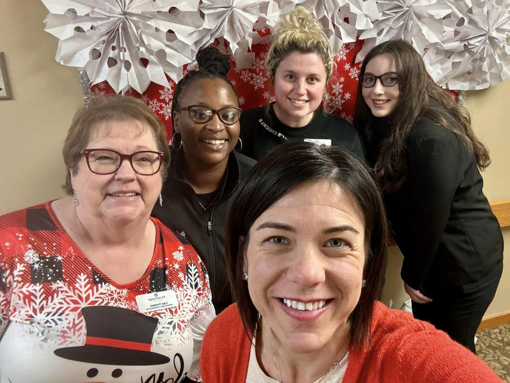Executive Director Karra and her team of employees are smiling in front of a decorated wall with large paper snowflakes and red wrapping paper.