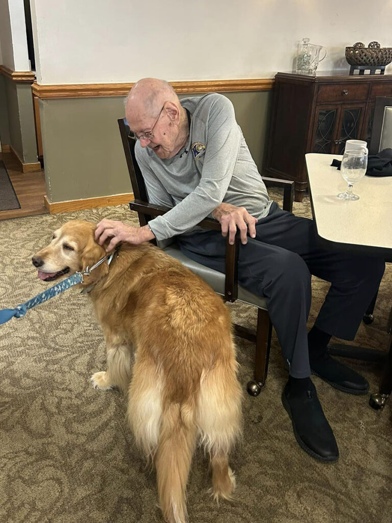Elderly man in chair petting a dog at senior living community canine comfort session.