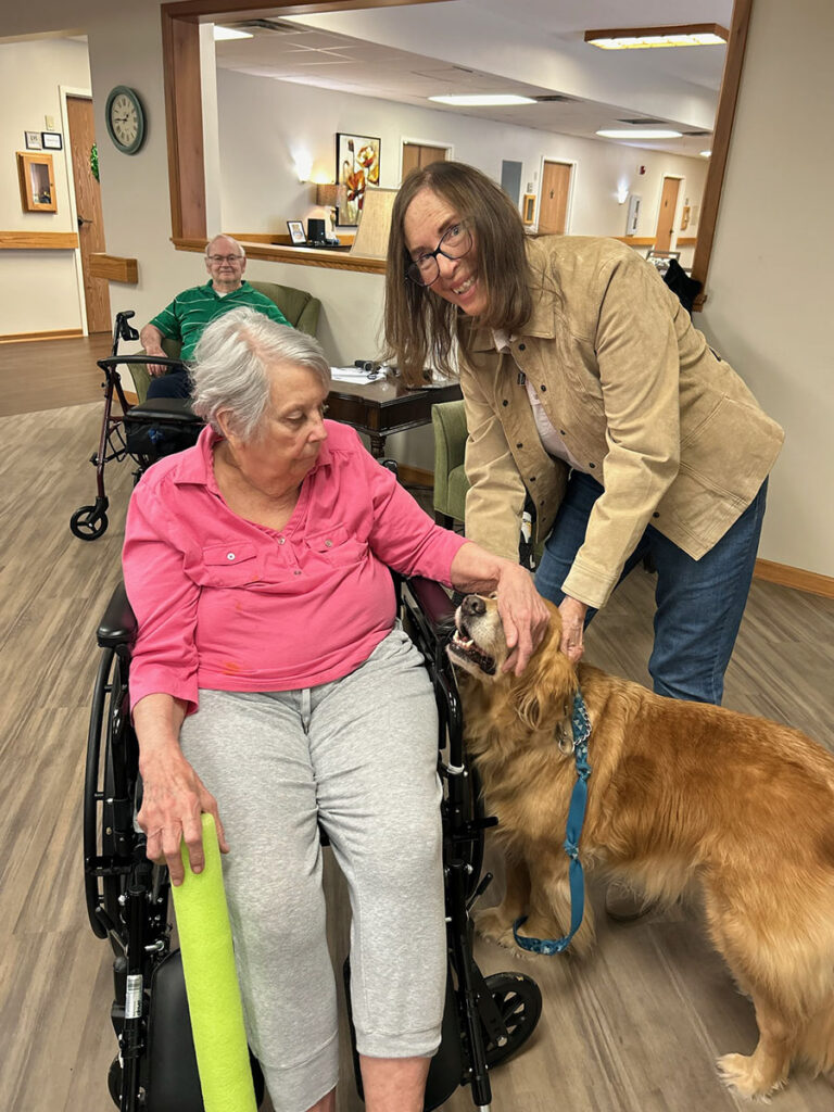 Spreading happiness, a volunteer brings a therapy dog to a memory care resident in a wheelchair, creating a heartwarming connection.