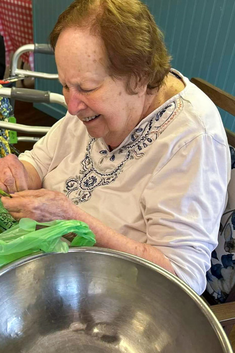 Smiling elderly woman sitting at a table, engaging in an activity with a green plastic bag and a large metal bowl at The Kentridge Senior Living.