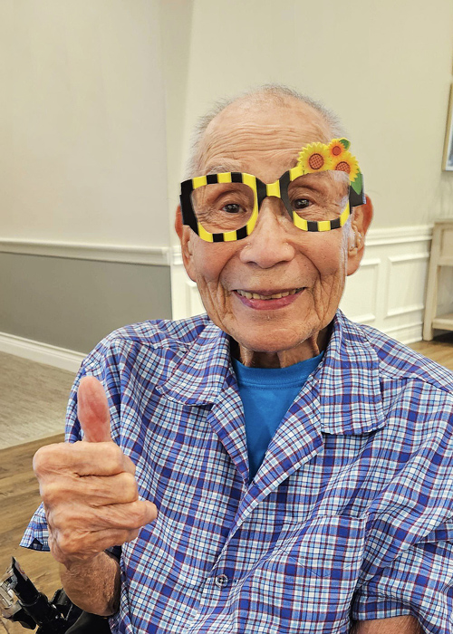 Senior resident giving a thumbs-up while wearing playful, sunflower-decorated glasses, radiating positivity, humor, and the joy of embracing life in a senior living community.