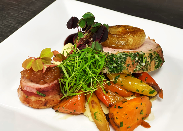 Gourmet plate featuring herb-crusted pork loin with roasted carrots and mashed potatoes, garnished with microgreens, accompanied by bacon-wrapped pork topped with chutney.