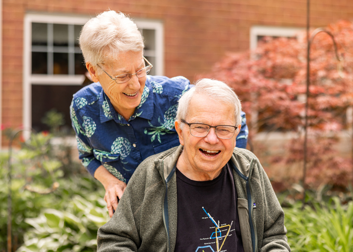 Happy senior resident sitting outdoors in a garden with vibrant plants, joyfully laughing with another resident standing behind him, both enjoying a lighthearted moment together.