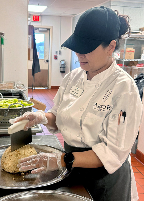 Chef in a white Arbor uniform and black cap using a bench scraper to portion a large ball of dough or cheese mixture in a professional kitchen.