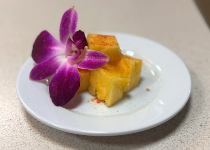White plate with cubed pineapple sprinkled with chili powder and garnished with a vibrant purple orchid, showcasing a sweet and spicy flavor pairing.