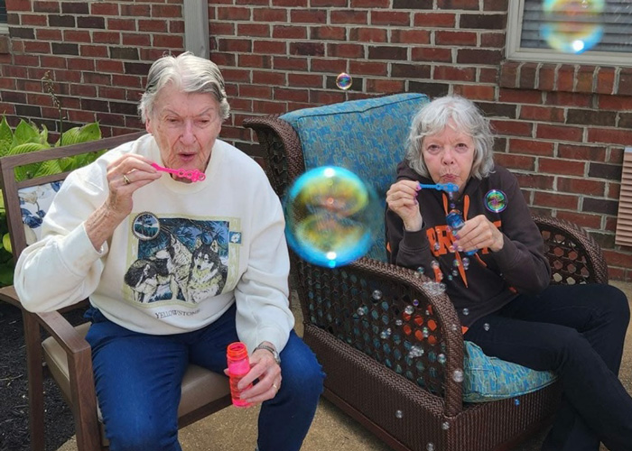 Two senior residents enjoy blowing bubbles while relaxing on a patio, surrounded by colorful floating bubbles.