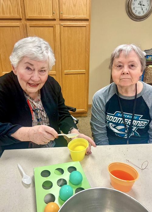 Two senior residents dye colorful Easter eggs at a table, smiling and enjoying the creative holiday activity in a warm and festive community environment.