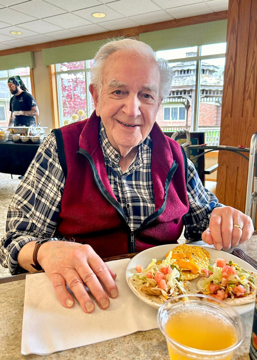Senior resident smiles while enjoying a festive Cinco de Mayo meal, featuring tacos and punch, with a lively community dining space in the background.