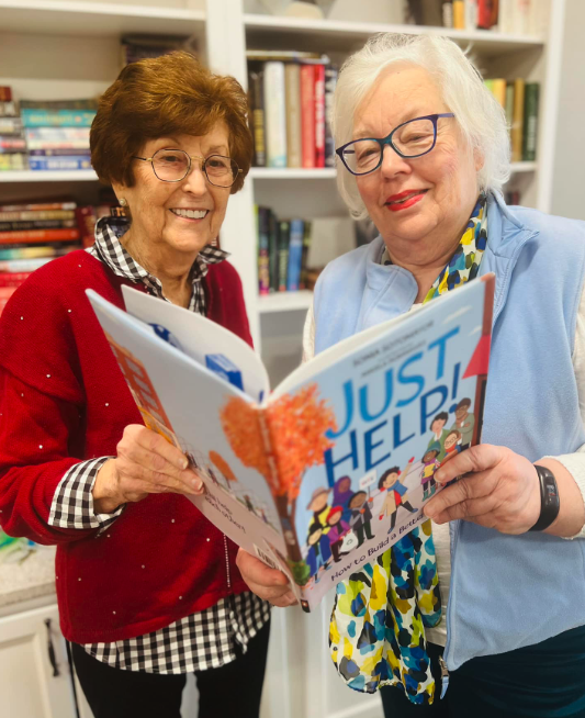 Two senior women smiling together while reading a book in a cozy library setting.