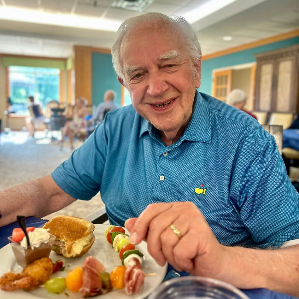 Smiling senior resident enjoys a fresh meal with skewers, fruit, bread, and dessert at the community.