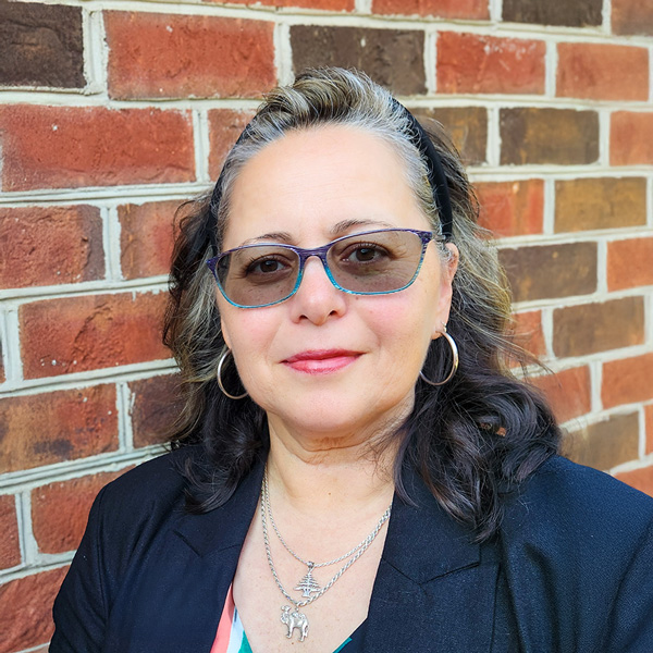 Gina Pratt, Senior Living Director at The Kentridge Senior Living, wearing glasses, hoop earrings, layered necklaces, and a black jacket, smiling in front of a brick wall.