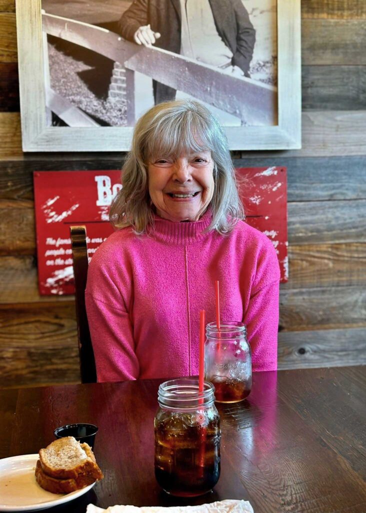 A cheerful resident in a bright pink sweater smiles at a restaurant table with mason jar drinks and a sandwich, enjoying a meal during a community outing.