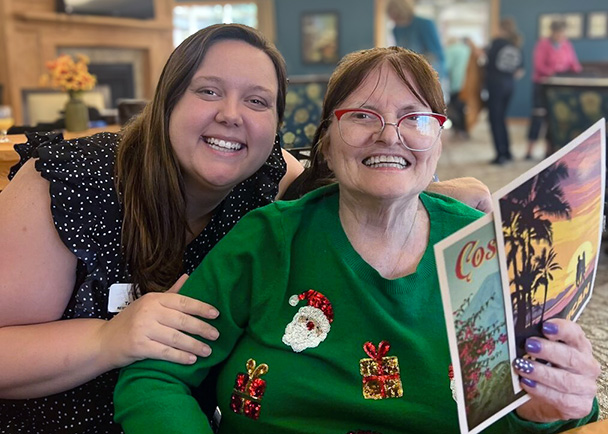 A team member and resident smile together, the resident wearing a festive Christmas sweater and holding travel related papers.