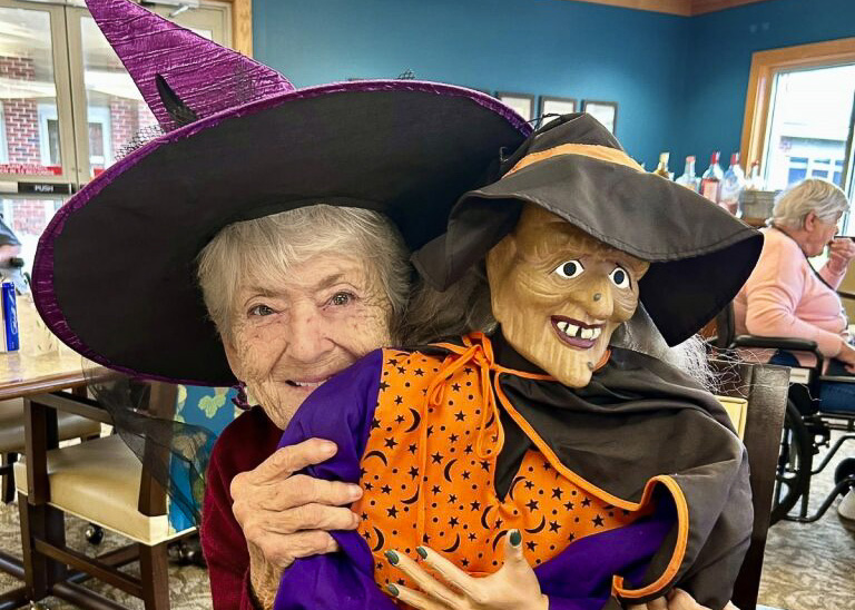 A senior woman wearing a witch's hat playfully holds a Halloween witch prop.