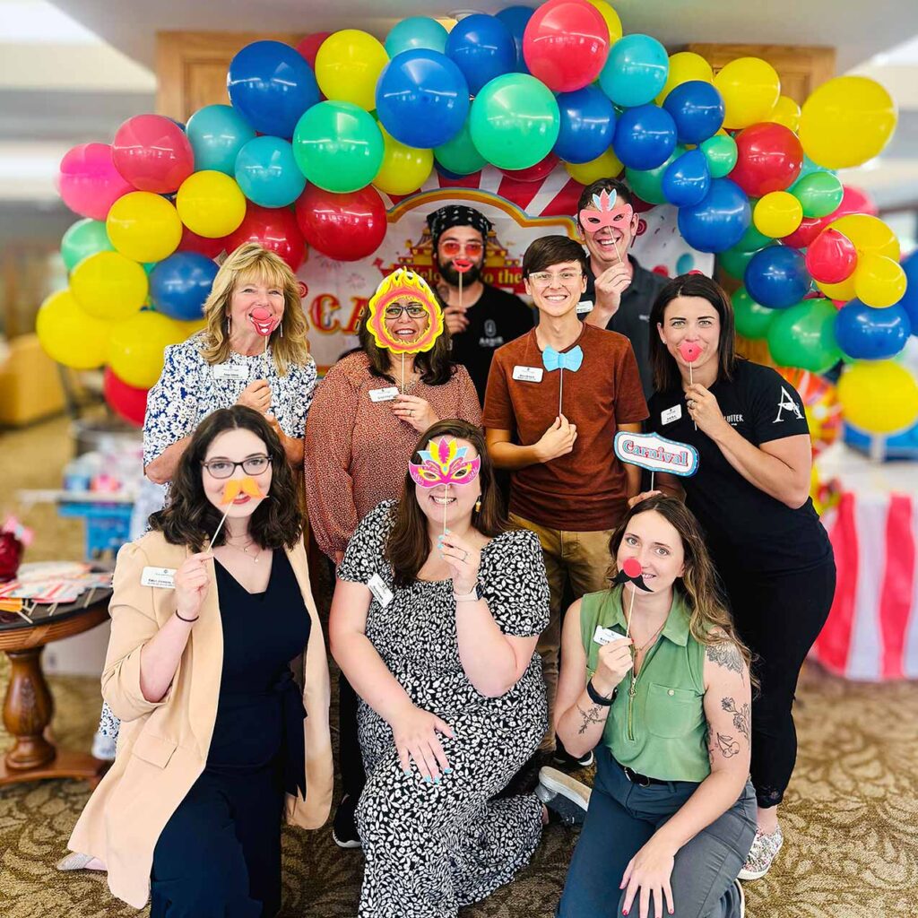 Team members at The KentRidge hold up novelty masks under a colorful balloon arch.