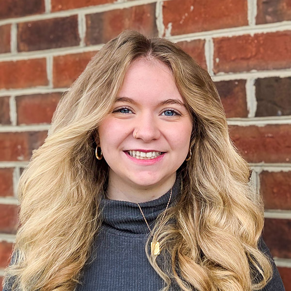 Ariel Jones, Resident Services Director at The Kentridge Senior Living, smiling in a gray top with long blonde hair, photographed against a red brick background.