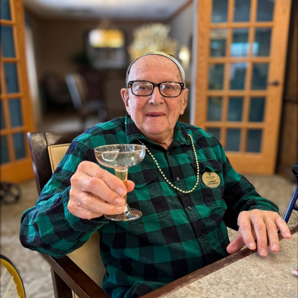 A senior resident smiles in a warm, inviting common space, raising a delicate glass in a toast.