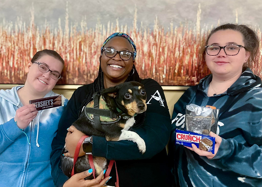 Three women on the Kentridge Senior Living team, one holding a small dog, smile brightly with sweet treats.