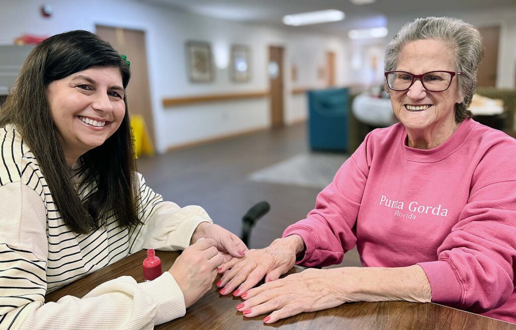 A loved one paints a resident's nails, both smiling brightly, during a relaxed day at The KentRidge Senior Living.