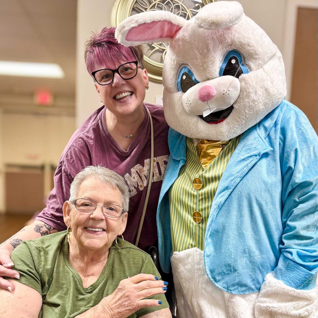 A senior woman, loved one, and Easter Bunny pose with bright smiles.