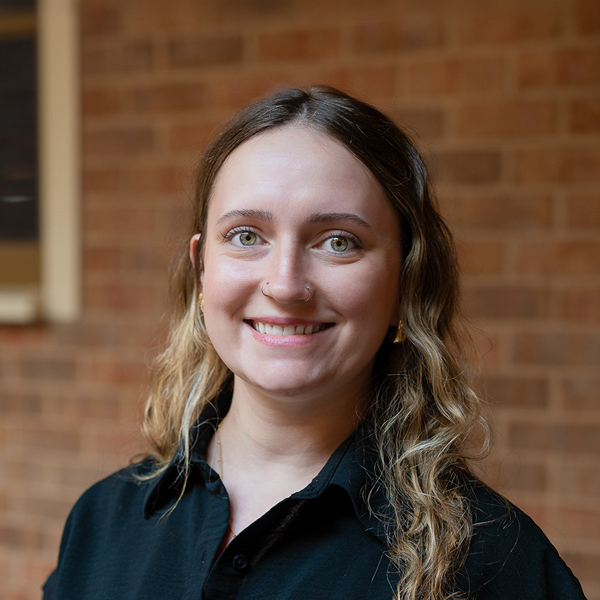 Alicia Miller, Wellness Director at The KentRidge Senior Living, smiling in a professional outdoor headshot with long wavy hair, small hoop earrings, a nose ring, a black collared top, and a softly blurred brick background.