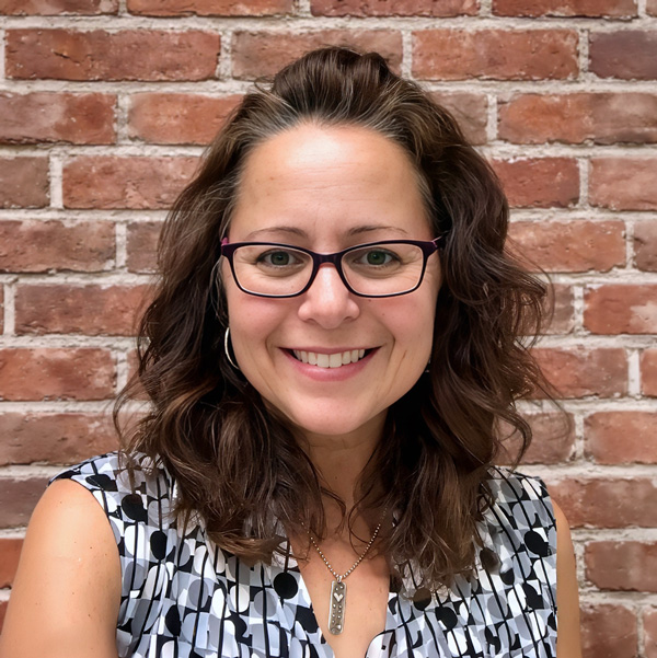 Gina Pratt, Senior Living Director at The Kentridge Senior Living, wearing glasses, printed black and white blouse, and a silver necklaces, smiling in front of a brick wall.