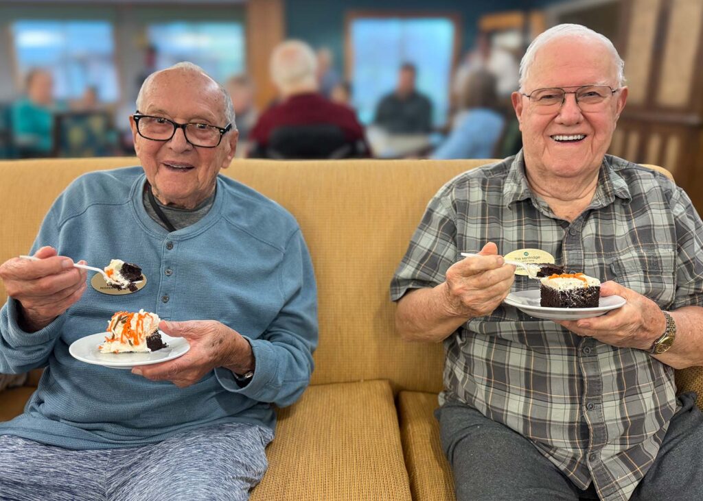 Two senior men at The Kentridge Senior Living smile brightly, enjoying desserts.