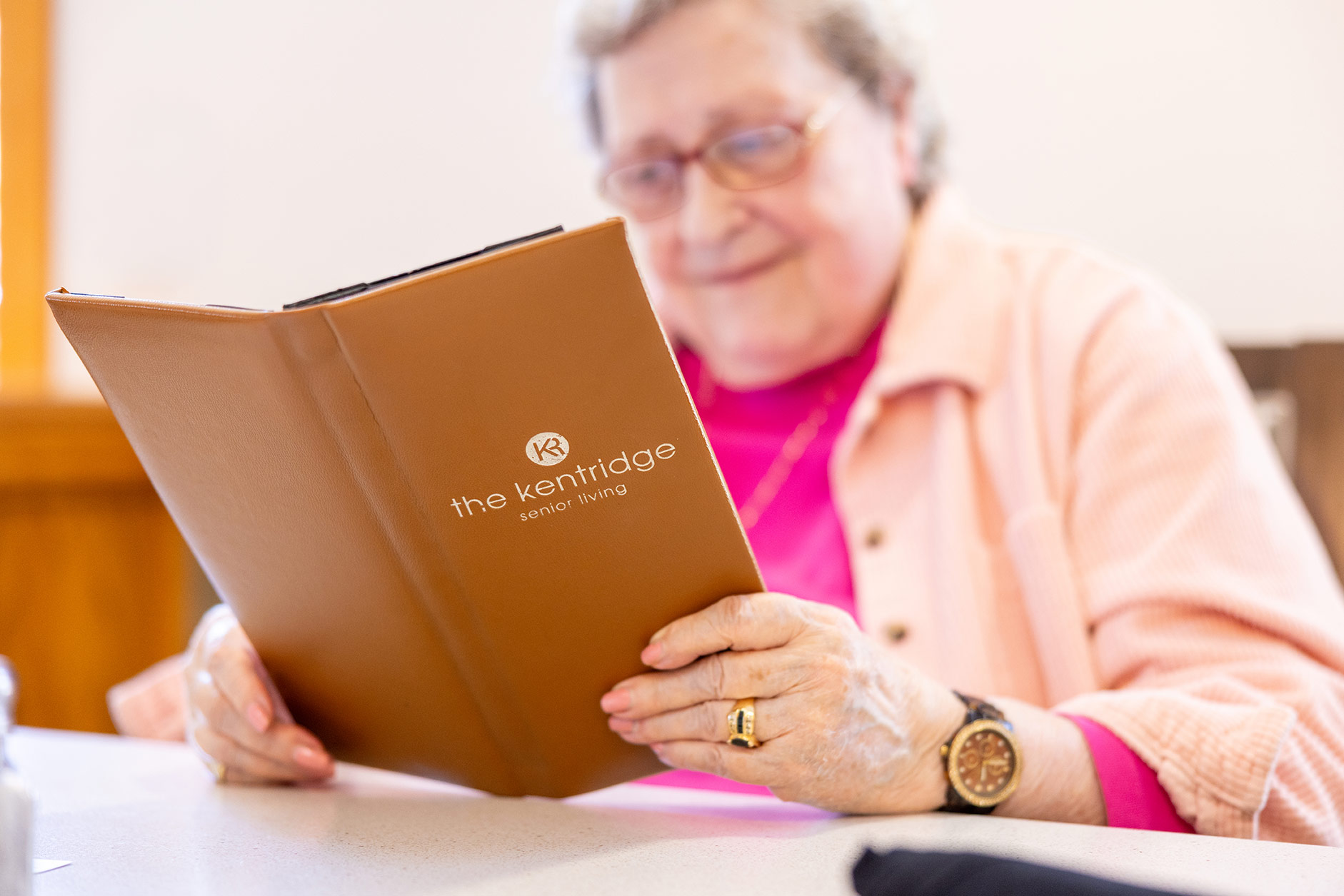 Resident smiling while reading a KentRidge Senior Living menu at a table in the community dining area.